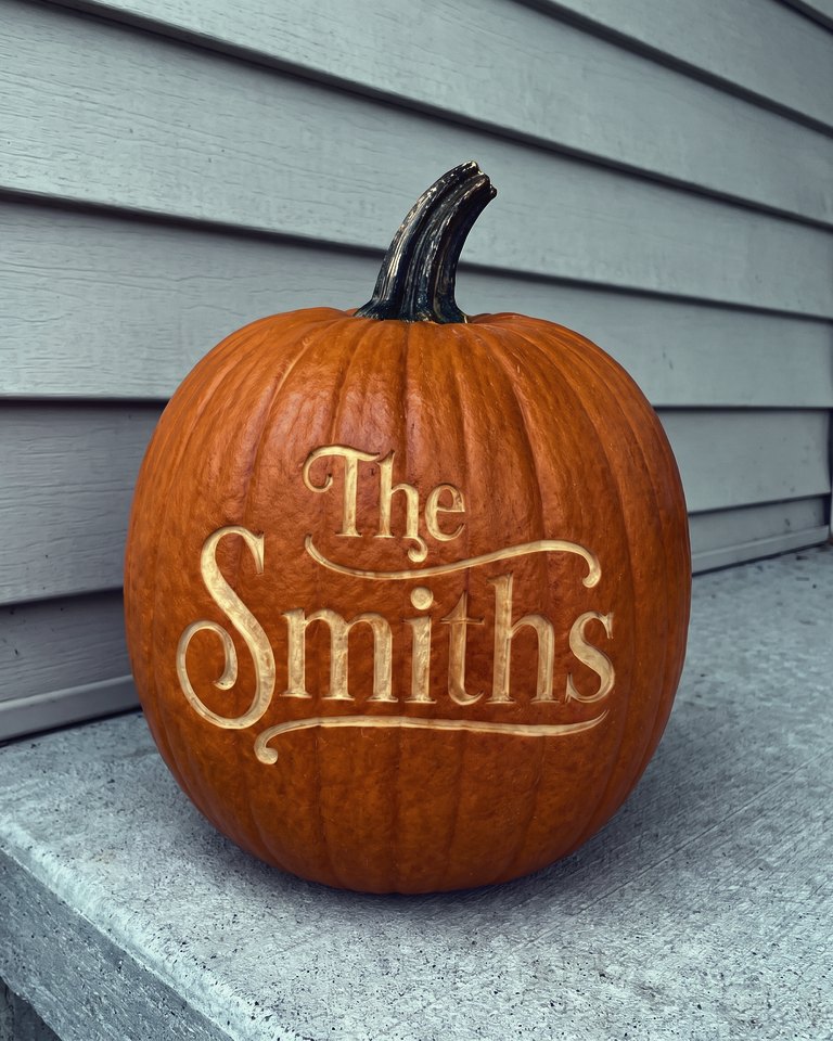 A pumpkin laser-engraved with the script words The Smiths, on a porch with mums